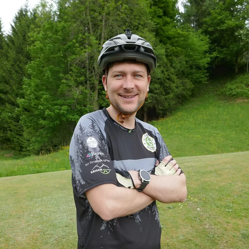 Mann mit Fahrradhelm und schwarzem Trikot steht l&auml;chelnd vor gr&uuml;ner Wiese und Wald.
