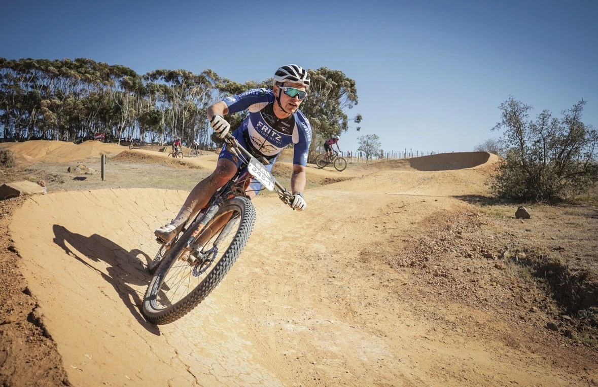 Mountainbiker in blauem Trikot f&auml;hrt &uuml;ber sandige Downhill-Strecke mit B&auml;umen im Hintergrund.