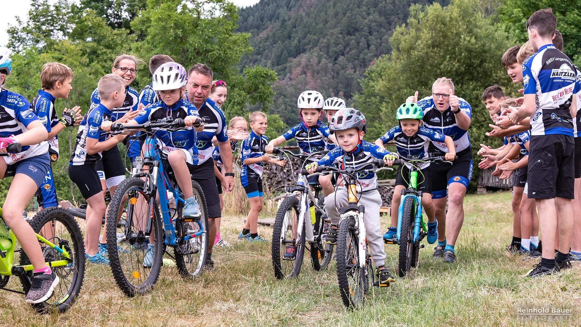Junge Radfahrer in blauen Trikots sind mit ihren Fahrr&auml;dern auf einer Wiese in bergiger Umgebung unterwegs.