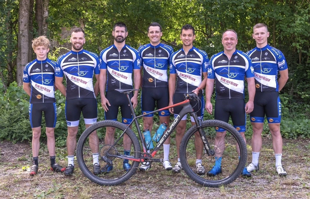 Sieben Mitglieder eines Rennradteams in blauwei&szlig;er Teamkleidung mit Fahrrad vor Baumkulisse.