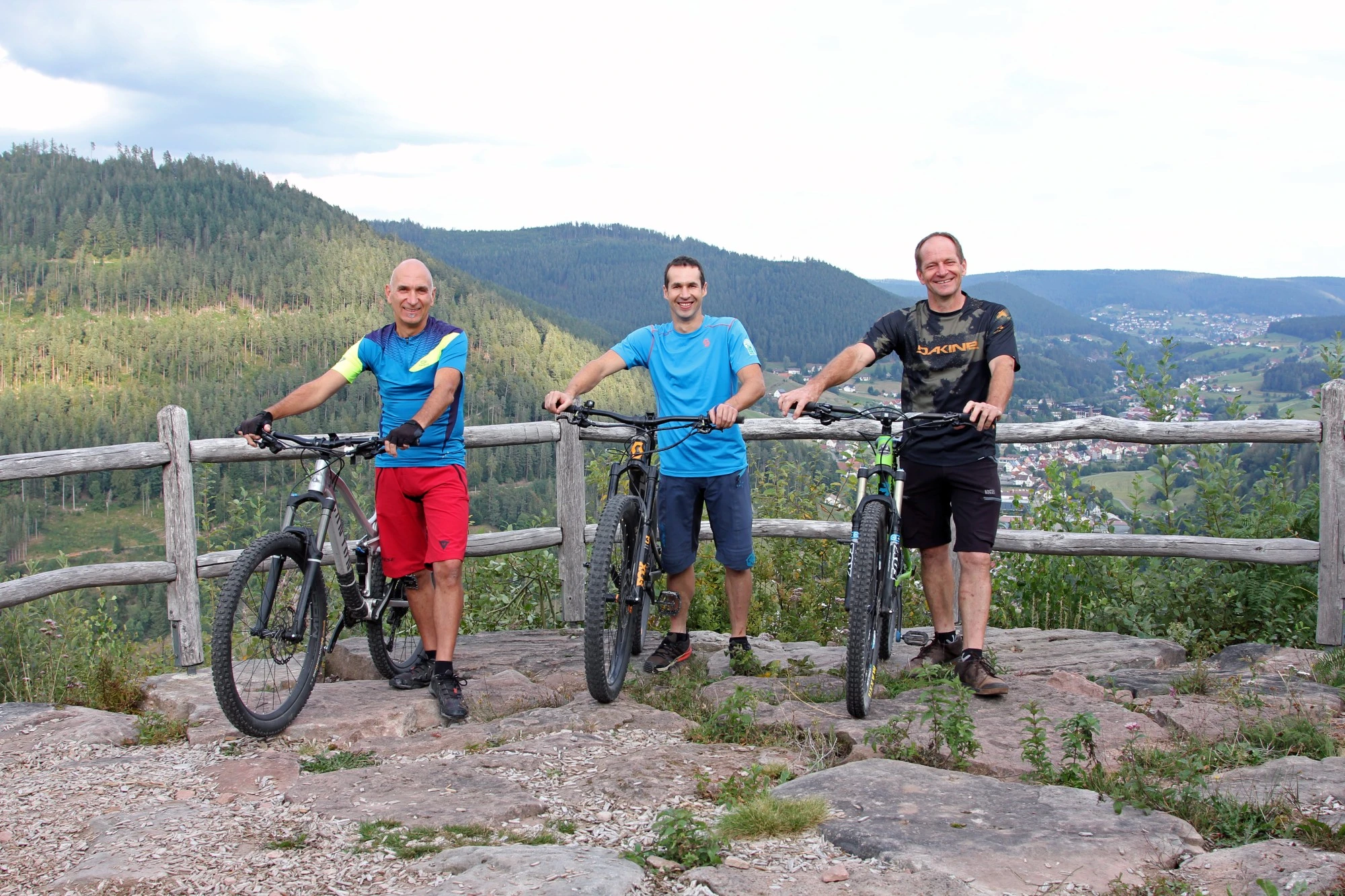 Drei Radfahrer mit Fahrr&auml;dern stehen an einer Aussichtsplattform mit Berglandschaft im Hintergrund.