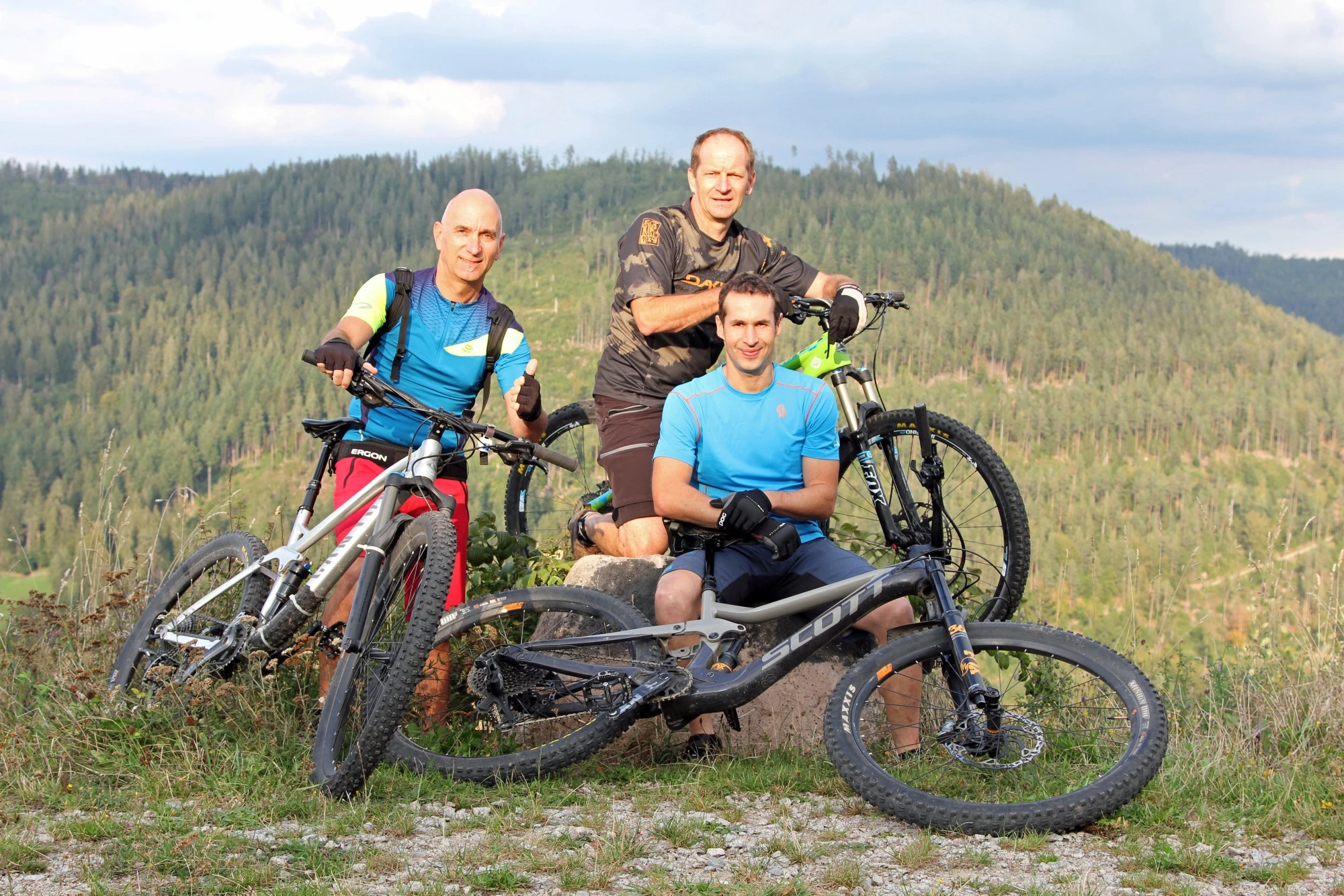 Drei Personen mit Mountainbikes in bergiger Waldlandschaft posieren f&uuml;r ein Gruppenfoto.