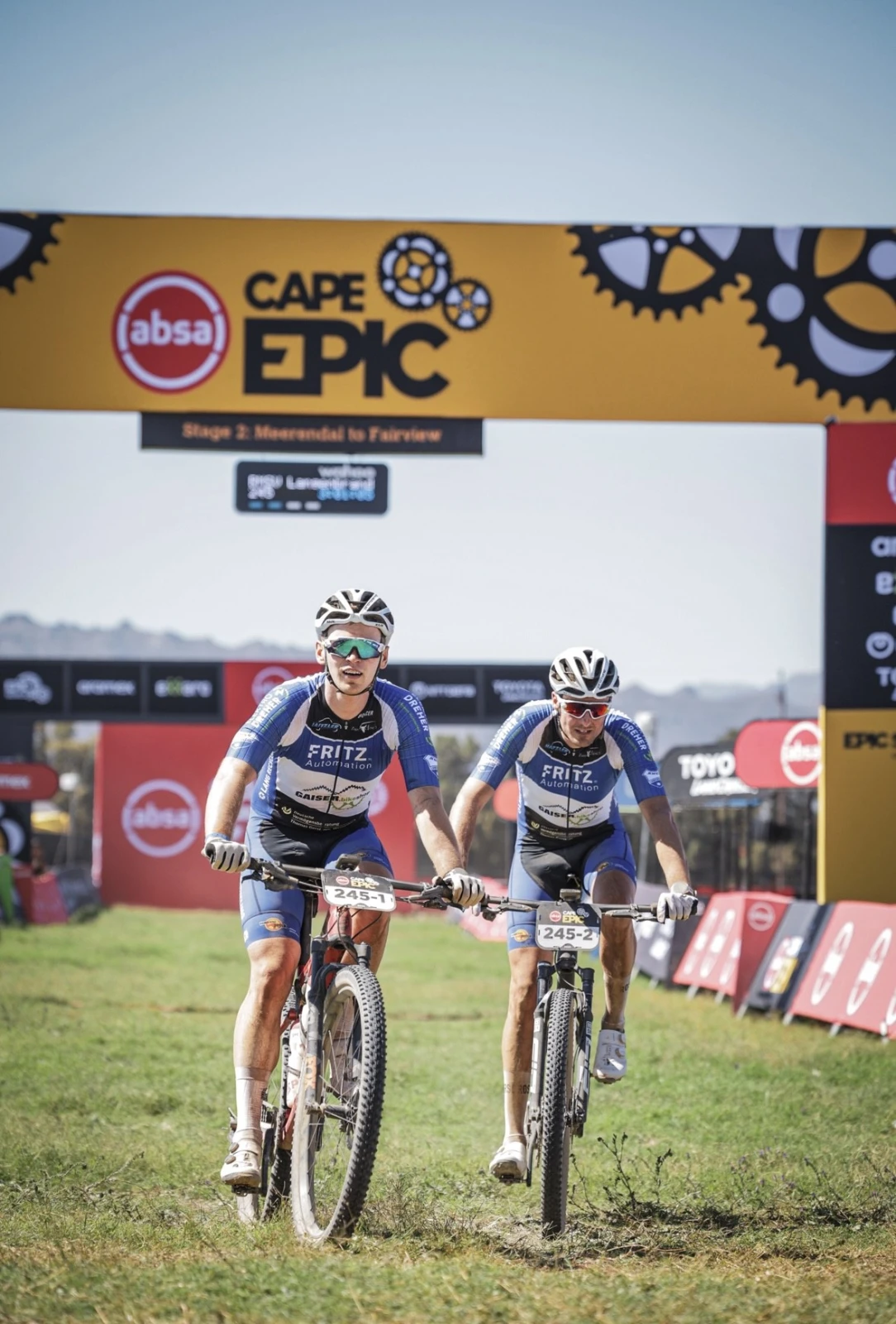 Zwei Mountainbiker im Team-Outfit fahren unter dem Cape Epic Zielportal &uuml;ber eine Wiese.