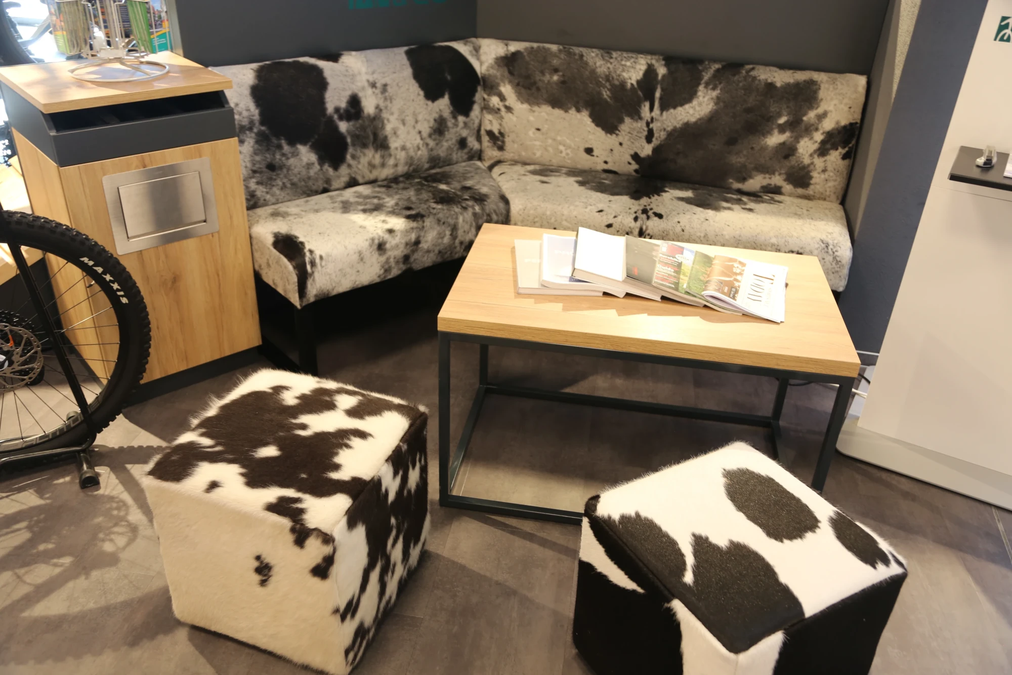 Gem&uuml;tliche Sitzecke mit grauem Sofa, Kuh-Mustern auf Poufs und Holztisch in einem Fahrradgesch&auml;ft.