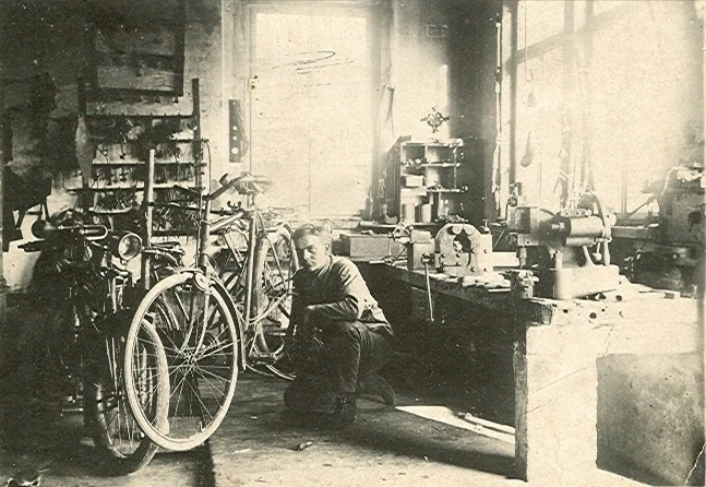 Historische Werkstatt mit Fahrr&auml;dern und Mechaniker bei der Arbeit an Arbeitspl&auml;tzen.